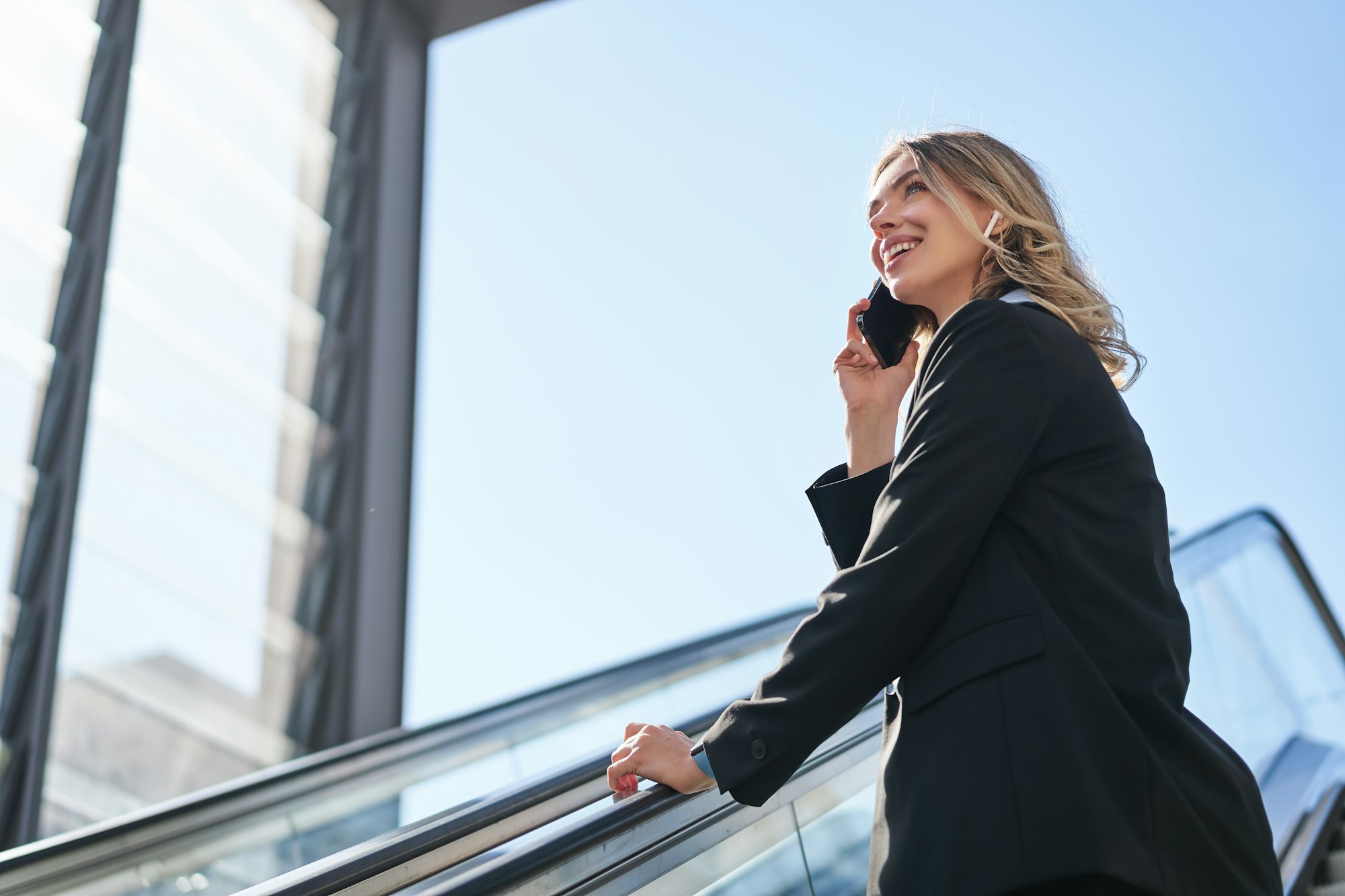 Portrait of businesswoman in black suit, going up on escalator, talking on mobile phone. Saleswoman